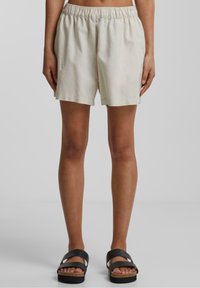 Shorts en lin beige clair avec une taille élastique, présentant une coupe décontractée et un design de jambe droite. Associés à des sandales noires à lanières.