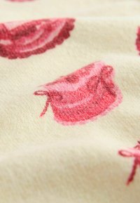 Baumwollstoff mit cremfarbenem Hintergrund, auf dem sich wiederholende rosa Cupcake-Illustrationen befinden, verziert mit gewellten Kanten und weicher Textur.