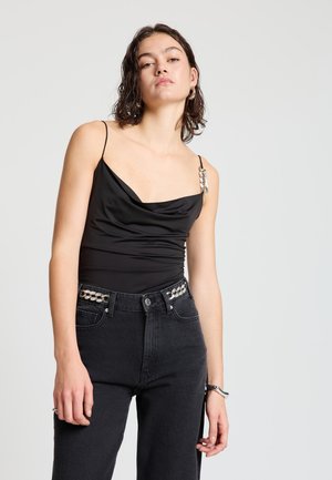 Black sleeveless top with draped neckline and silver hardware detail on one strap, paired with high-waisted black jeans with a patterned belt.