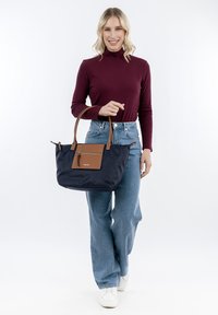 Marine blauwe tote bag met bruine leren accenten, twee stevige handvatten, een ritsvak aan de voorkant en een ruime binnenkant, gedragen door een model.