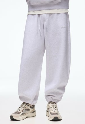 Løse lysegrå joggingbukser med elastiske manchetter båret sammen med beige og grå sneakers på en person, der står mod en hvid baggrund.