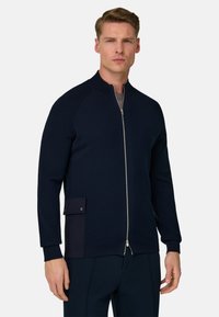 Chaqueta de punto azul marino con cremallera, cuello acanalado y bolsillo lateral. Presenta una textura suave y un diseño ajustado, combinada con pantalones oscuros.