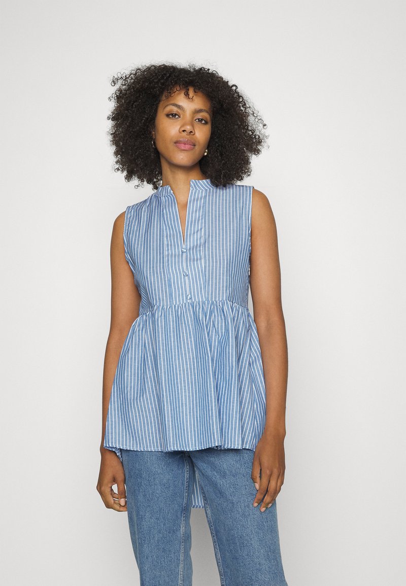 Blusa sem mangas azul às riscas com uma pala de botões, cintura franzida e decote alto. Combinada com calças de denim azul claro.
