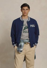 Jonge man in een marineblauwe varsityjack over een grijze graphic tee, beige broek en een geruit overhemd om zijn middel geknoopt, staande met zijn handen in zijn zakken.