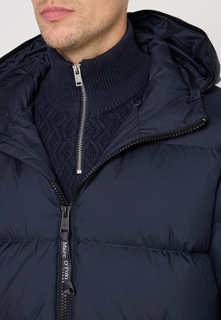 Mann in einer navyblauen gepolsterten Jacke mit Kapuze über einem navyblauen, kabelgestreiften Pullover mit einem silbernen Halbzip-Kragen.