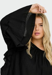 Crna bluzica s naboranim, lepršavim rukavima. Lagani, teksturirani materijal s suptilnim sjajem. Nabrani izrez i meki obrisi.