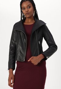 Crna kožna jakna s širokim ovratnikom, patentnim zatvaračem i skraćenim dizajnom, u kombinaciji s rebrastom burgundskom haljinom.