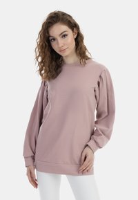 Felpa rosa a maniche lunghe realizzata in tessuto morbido, con maniche a sbuffo e scollo rotondo, abbinata a pantaloni bianchi attillati.