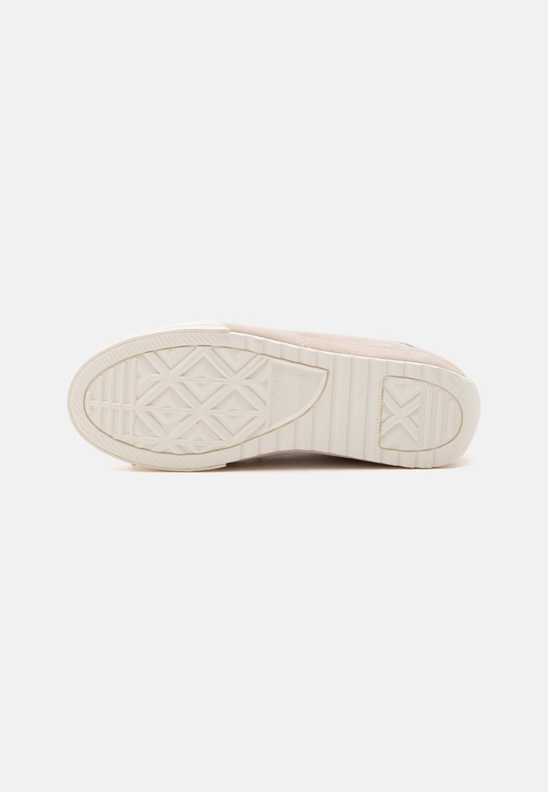 Flache beige Schuhsohle mit geometrischen Profilmustern und einem kleinen runden Logo nahe der Ferse auf einfachem hellem Hintergrund.