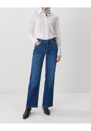 Person iført en hvid langærmet skjorte, mønstret halstørklæde, højtaljede blå viddeben jeans og sorte hælede ankelstøvler.