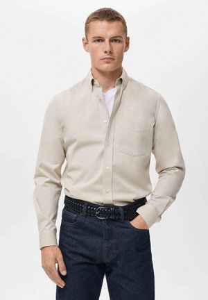 Man wearing beige button-up shirt over white tee, dark blue jeans with braided black belt, hand in pocket, neutral expression.