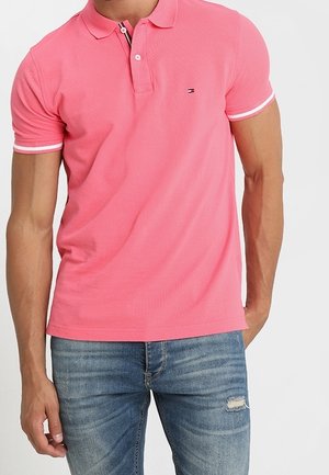 Polo rose avec une surface texturée, manches courtes, patte à deux boutons et rayures d'accent blanches. Porté avec un jean bleu déchiré.