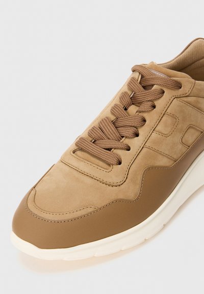 Sneaker da uomo color beige con upper in suede, dettagli pannellati cuciti, lacci marroni e suola bianca su sfondo bianco.