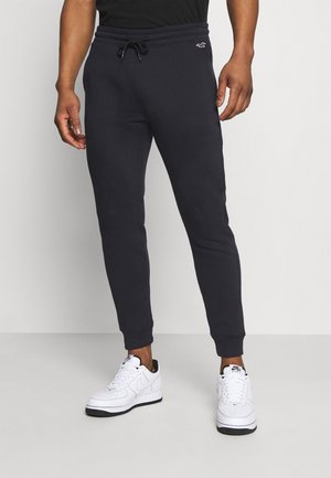 Mężczyzna w czarnych, dopasowanych spodniach dresowych z sznurkiem w pasie oraz białych butach Nike Air Force 1, stojący na jednolitym szarym tle.