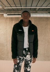 Schwarze und graue Jacke mit Fleece-Futter und gepolsterten Abschnitten, ausgestattet mit einem Reißverschluss und gestickten Logos, kombiniert mit schwarz-weißen gemusterten Hosen.