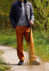 Padded black jacket, gray t-shirt, and orange pants on a person holding a trekking pole, standing on a path surrounded by greenery.