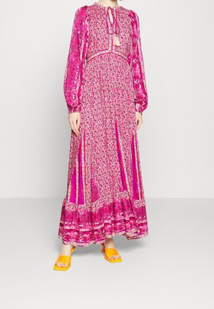 Kobieta w długiej różowej sukience w kwiaty z długimi rękawami i ozdobnym wiązaniem przy szyi, w połączeniu z jasnoniebieskimi sandałami na obcasie z odkrytymi palcami.