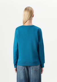 Teal-coloured collegepaita, jossa on pitkät hihat ja pyöreä pääntie. Paidan helmassa ja hihoissa on resorit, ja se on yhdistetty löysiin sinisiin farkkuihin.
