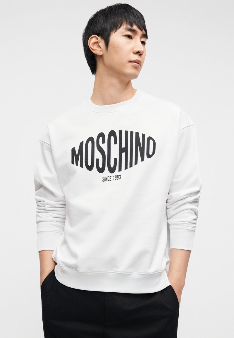 Sweat-shirt blanc avec un grand logo noir "MOSCHINO" et le texte "SINCE 1983". Il possède un col rond et des poignets côtelés. Design simple et moderne.