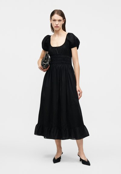 Γυναίκα στέκεται κρατώντας μια μαύρη τσάντα, φοράει μαύρο midi φόρεμα με φουσκωτά μανίκια και βολάν στο κάτω μέρος, και μαύρες μυτερές γόβες.