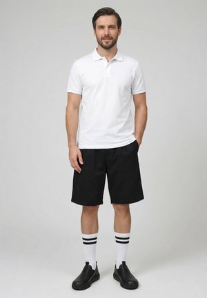 Mann stehend, trägt weißes Poloshirt, schwarze knielange Shorts, weiße Socken mit schwarzen Streifen und schwarze Slipper, neutraler grauer Hintergrund.