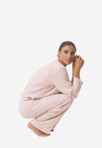 Roze geribbelde loungewear set met een relaxte pasvorm, met lange mouwen en wijde broekspijpen, getoond in een gehurkte positie tegen een witte achtergrond.