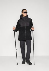 Melna fleece jaka ar kviltotu melnu augšējo paneli; saskaņota ar tumšām biksēm un pārgājienu zābakiem. Modelis tur pārgājienu nūjas un valkā saulesbrilles.