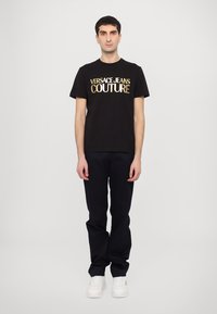 Juodas medvilninis marškinėliai su auksiniu "VERSACE JEANS COUTURE" logotipu, derinami su tamsiomis kelnėmis ir baltais batais, stovint neutraliame fone.