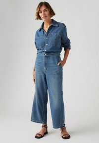 Jumpsuit en denim bleu clair avec des manches longues, deux poches poitrine, une taille élastique, une coupe large et une fermeture à boutons.