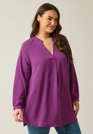 Maglia lunga viola con design a scollo a V, tessuto texture e polsini arricciati. Abbinata a jeans blu, che mostrano una vestibilità comoda.