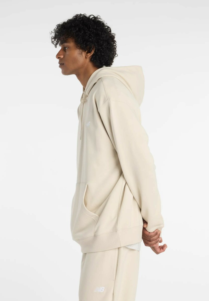 Beige Kapuzenpullover mit einer vorderen Tasche und Kordelzug-Kapuze, mit einem kleinen Logo auf der Brust, aus einem weichen, strukturierten Material.