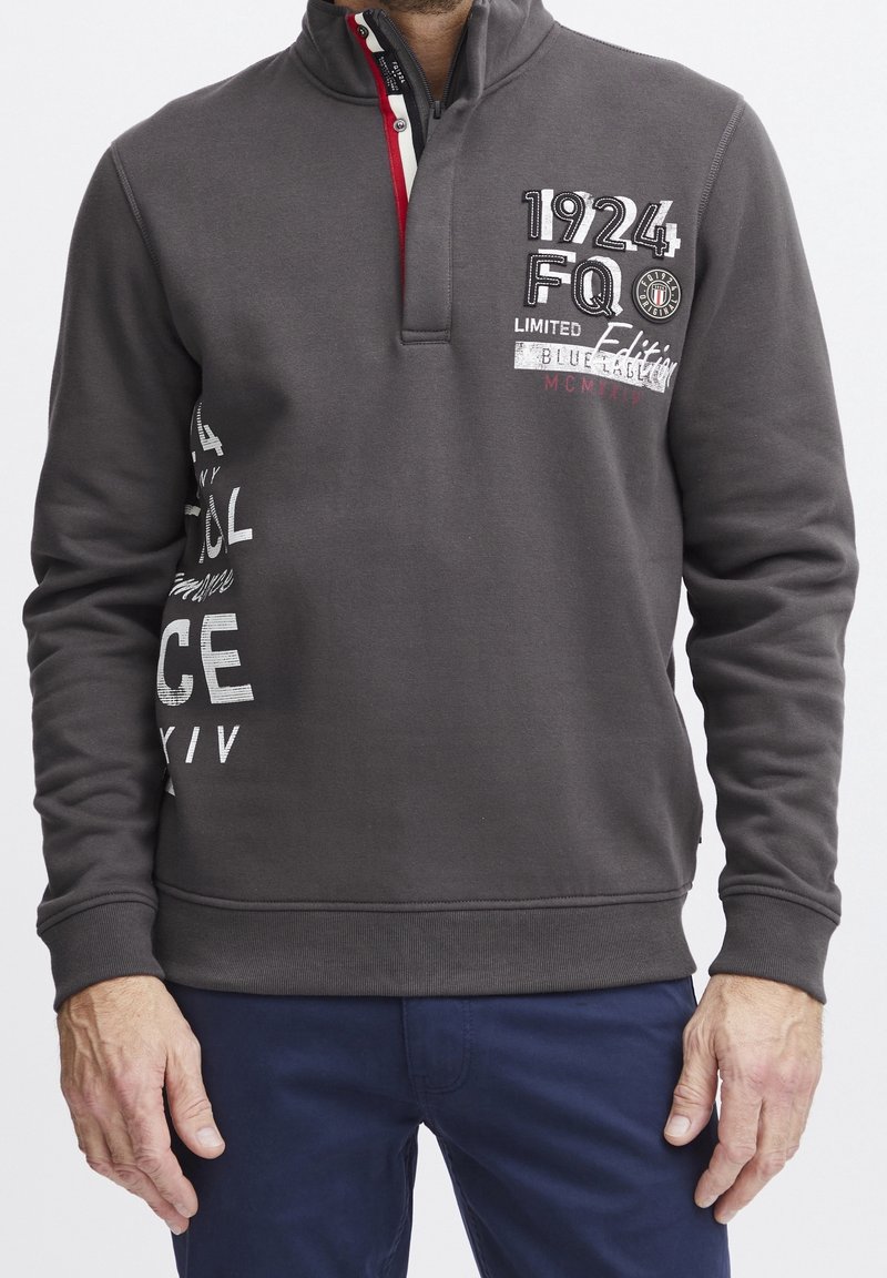 Homme portant un pull gris foncé avec une fermeture éclair au quart et un texte imprimé, associé à un pantalon bleu foncé, debout sur un fond blanc.