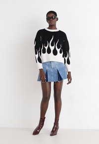 Μαύρος και λευκός πουλόβερ με σχέδιο φλόγας και στρογγυλή λαιμόκοψη, συνδυασμένο με πλισέ τζιν φούστα και μποτάκια μπορντό μέχρι τον αστράγαλο. Φοριούνται γυαλιά ηλίου.