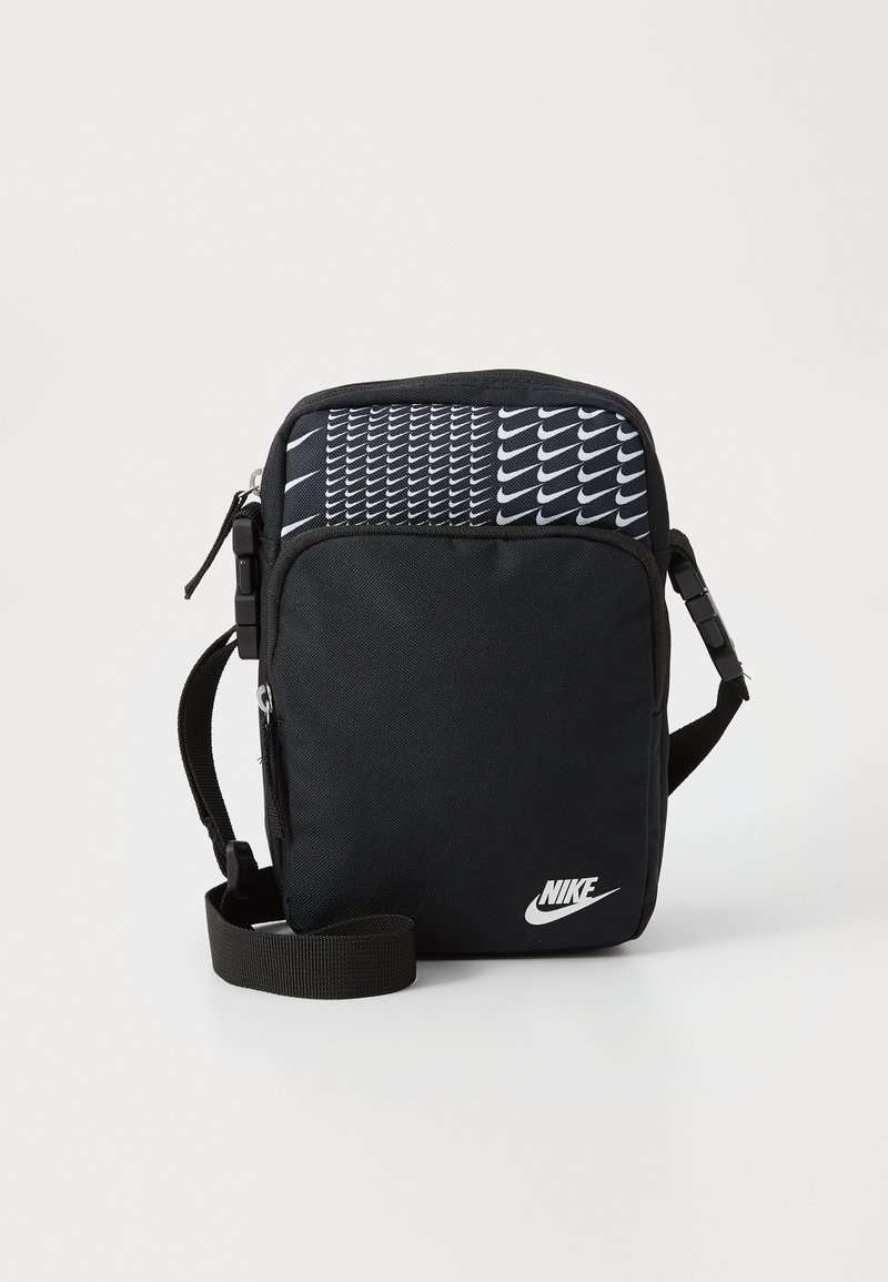 Bolso cruzado de tela negra con un patrón de cuadrícula de logotipos blancos de Nike, cierre superior con cremallera, correa ajustable y logotipo destacado en el frente.