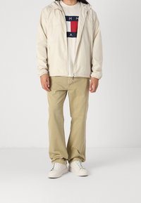 Světle béžová bunda s kapucí, zapínání na zip, elastické manžety; světle hnědé kalhoty; bílé tenisky; tričko s potiskem v námořnické modři a červené barvě.