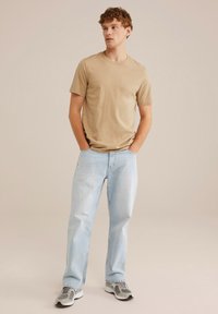 Camiseta de algodón beige de mangas cortas, combinada con jeans de denim azul claro. Modelo con zapatillas grises y blancas, de pie con las manos en los bolsillos.