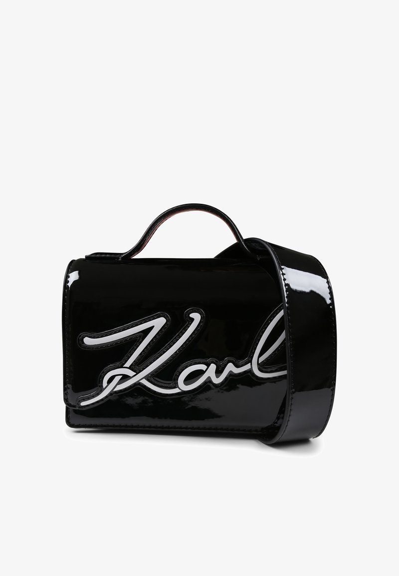 Sac à main en cuir verni noir avec un logo "Karl" blanc bien en évidence, forme structurée, anses supérieures et bandoulière amovible.