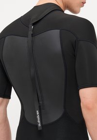 Fekete neoprémből készült rövid wetsuit, sima hátrésze, cipzáras záródása és varrott díszítése van. A márka logója a hát alsó részén látható.