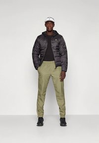 Juoda pūkinė striukė su gaubtu, žalios kelnės ir juodi sportbačiai. Vyras stovi tiesiai, dėvi juodą marškinėl�į ir baltą kepuraitę su logotipu.