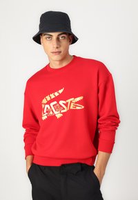 Felpa rossa con logo dorato Lacoste, polsini e scollatura a coste. Indossata con pantaloni neri e un cappello a secchiello blu navy. Sfondo semplice.