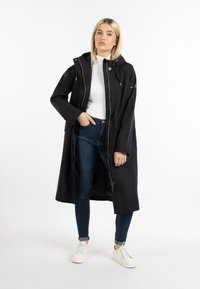 Schwarzer wasserfester langer Mantel mit Kapuze, mit Knöpfen und Reißverschluss. Getragen über einem weißen Oberteil, kombiniert mit blauen Jeans und weißen Turnschuhen.