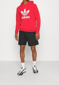 Roter Kapuzenpullover mit weißem Adidas-Logo, schwarze Shorts und schwarz-weiße Sneaker. Das Outfit zeichnet sich durch einen lässigen, sportlichen Stil mit lockerer Passform aus.