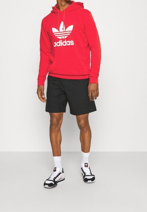 Czerwona bluza z kapturem z białym logo Adidas, czarne szorty oraz czarno-białe sneakersy. Strój charakteryzuje się swobodnym, sportowym stylem z luźnym krojem.