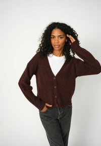 Cardigan bordeaux lavorato a maglia con scollatura a V e cinque bottoni, caratterizzato da maniche lunghe e una vestibilità rilassata, abbinato a jeans scuri.