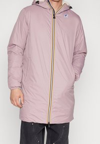 Homme portant une longue veste à capuche de couleur violet clair avec une fermeture éclair rayée et un logo sur la poitrine, les mains dans les poches, associée à un pantalon foncé.