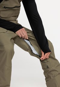 Olijfgroene overall met een ritssluiting bij de enkel, elastische taille en zwarte gestructureerde binnenvoering. Bevat een klein logo op het been.