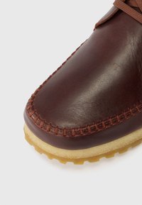 Sapato de couro castanho com costura visível, bico arredondado e sola de borracha de cor clara. A textura é suave com um acabamento mate.