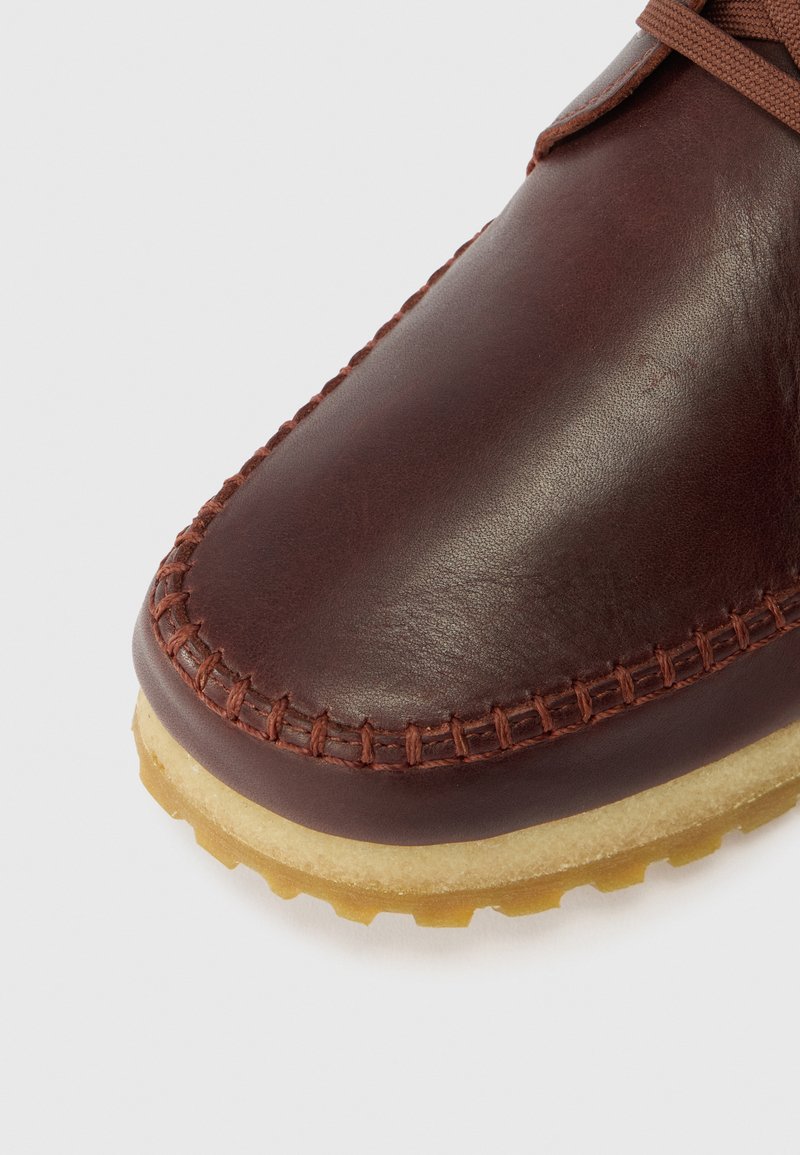 Braunlederner Schuh mit sichtbaren Nähten, runder Zehenpartie und einer hellen Gummisohle. Die Oberfläche ist glatt mit einem matten Finish.