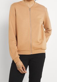 Modelo vistiendo una chaqueta de cremallera color marrón claro, con un primer plano de manos tatuadas, sobre un fondo blanco liso.