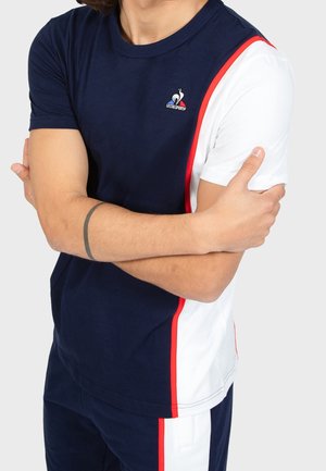 Homme portant un t-shirt Le Coq Sportif bleu marine et blanc avec des bordures rouges, les bras croisés, tatouage visible sur l'avant-bras gauche.